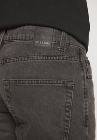 Vista trasera de una persona que lleva jeans negros desgastados con una etiqueta de la marca "Only & Sons" en la cinturilla y una camisa negra.
