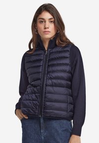 Mörkblå puffväst med dragkedja framtill, med quiltade sektioner, hög krage och ärmar i mörkblått stickat, i kombination med jeans.