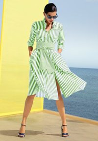 Gestreiftes Hemdkleid in Grün und Weiß, mit tailliertem Bund, langen Ärmeln und einem Schlitz bis zur Mitte des Oberschenkels. Hat einen Kragen und Taschen.