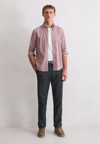 Casualny strój z jasnoróżową koszulą zapinaną na guziki, białym t-shirtem, ciemnoszarymi spodniami i beżowymi zamszowymi butami. Prosty design i neutralne kolory.