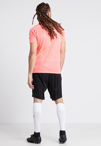 Vyriškas sportinis outfit'as, apimantis ryškiai rožinę trumpą rankovę marškinėlį, juodas šortus, baltas kelio ilgio kojines ir juodus sportinius batelius.