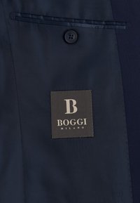 Intérieur d'un costume bleu marine foncé montrant un bouton noir au-dessus d'une étiquette cousue avec le texte "B Boggi Milano" en blanc.