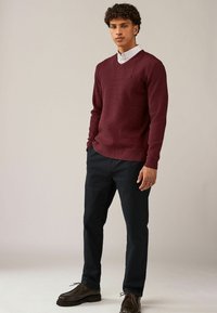 Sweater en V burgundy en tissu texturé, associé à un pantalon bleu marine foncé et des chaussures marron à lacets, se tenant devant un arrière-plan neutre.