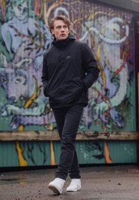 Czarna wodoodporna kurtka z kapturem, z przodu na zamek błyskawiczny i bocznymi kieszeniami. Połączona z ciemnymi dżinsami i białymi trampkami, na tle kolorowych graffiti.