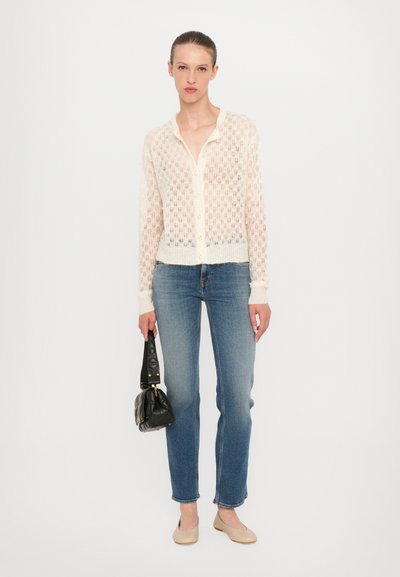 Strickjacke in hellem Creme mit einem Rautenmuster, geknöpfte Vorderseite und langen Ärmeln, kombiniert mit blauen Jeans und einer kleinen schwarzen Handtasche.