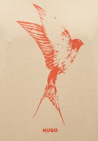 Κόκκινο στυλιζαρισμένο πουλί σε πτήση με ανοιχτά φτερά σε μπεζ φόντο, με το κείμενο "HUGO" από κάτω.
