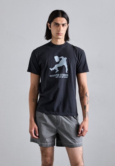 Black cotton t-shirt featuring a graphic print in light blue, with the text “KIDSUPER STUDIOS” and an illustration of a skateboarder. Worn with gray shorts.
