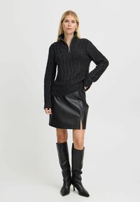 Schwarzer Strickpullover mit hohem Kragen und Reißverschluss, kombiniert mit einem schwarzen Leder-Minikleid mit seitlichem Schlitz und kniehohen schwarzen Stiefeln.