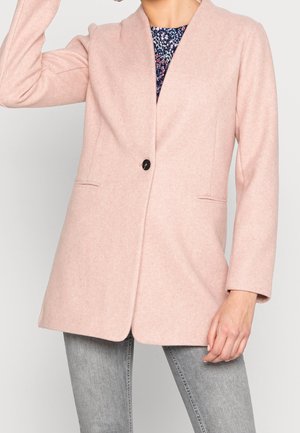 Vrouw met een lichtroze enkelgespakte blazer over een patroon top en grijze jeans, met één arm omhoog en de andere rustend.