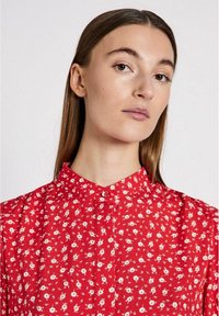Blusa rossa con colletto, caratterizzata da un motivo floreale con fiori bianchi e gialli. Realizzata in tessuto leggero, con chiusura a bottoni frontale e leggere arricciature sulle maniche.