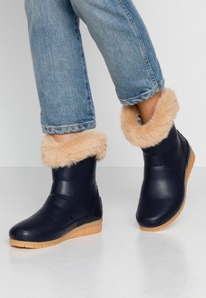 Kobieta nosząca niebieskie botki z beżowym futrzanym obramowaniem i jasnoniebieskie dżinsy na jednolitym jasnym tle.