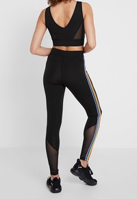 Soutien-gorge de sport noir avec un dos en V, associé à un legging noir avec des rayures au couleur arc-en-ciel sur les côtés et des panneaux en maille transparente sur les jambes. Baskets noires.