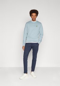 Felpa azzurra chiara con colletto tondo e piccolo logo giallo, abbinata a pantaloni blu scuro e sneakers bianche, con una texture liscia.