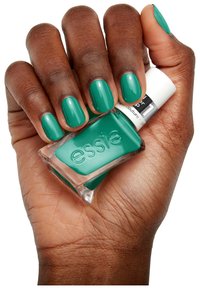 Groene nagellak in een heldere glazen fles met gegraveerd merk, vastgehouden in een hand met verzorgde nagels, ook groen geverfd.