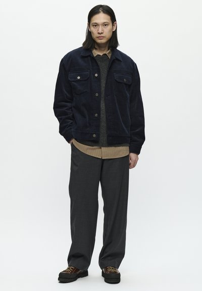 Námořnická kordová bunda s dvěma náprsními kapsami přes texturovaný šedý svetr a béžovou košili s límečkem, doplněná šedými kalhotami a hnědými botami.