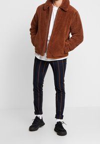 Brown fleece jacket with a collar, zip closure, paired with navy striped pants featuring brown vertical lines and black sneakers.