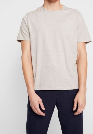 Man wearing light beige short-sleeve T-shirt with chest pocket and dark navy pants, standing against a plain light background.