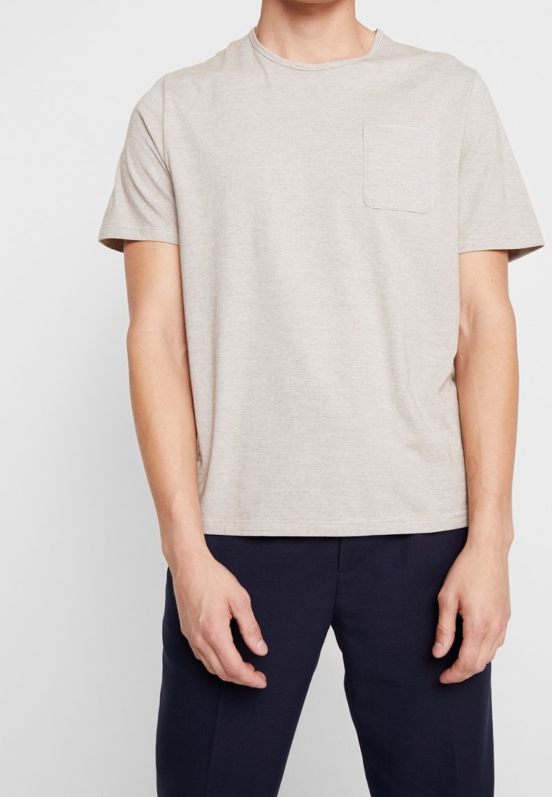 Homme portant un t-shirt à manches courtes beige clair avec une poche poitrine et un pantalon bleu marine foncé, debout devant un fond clair uni.