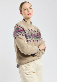 Beige trui met kleurrijke geometrische patronen in paars en groen, ronde halslijn, textuur van wollen stof, gecombineerd met lichte broeken.