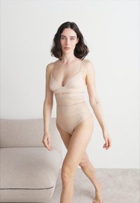 Huidkleurige bodysuit met dunne bandjes, naadloos ontwerp en een strakke pasvorm. Zachte textuur, gladde stof en minimalistische stijl.