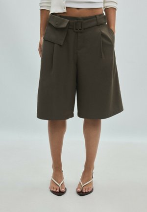 Femme portant un short vert foncé ceinturé aux genoux avec une poche à rabat et des sandales blanches, debout devant un fond clair uni.