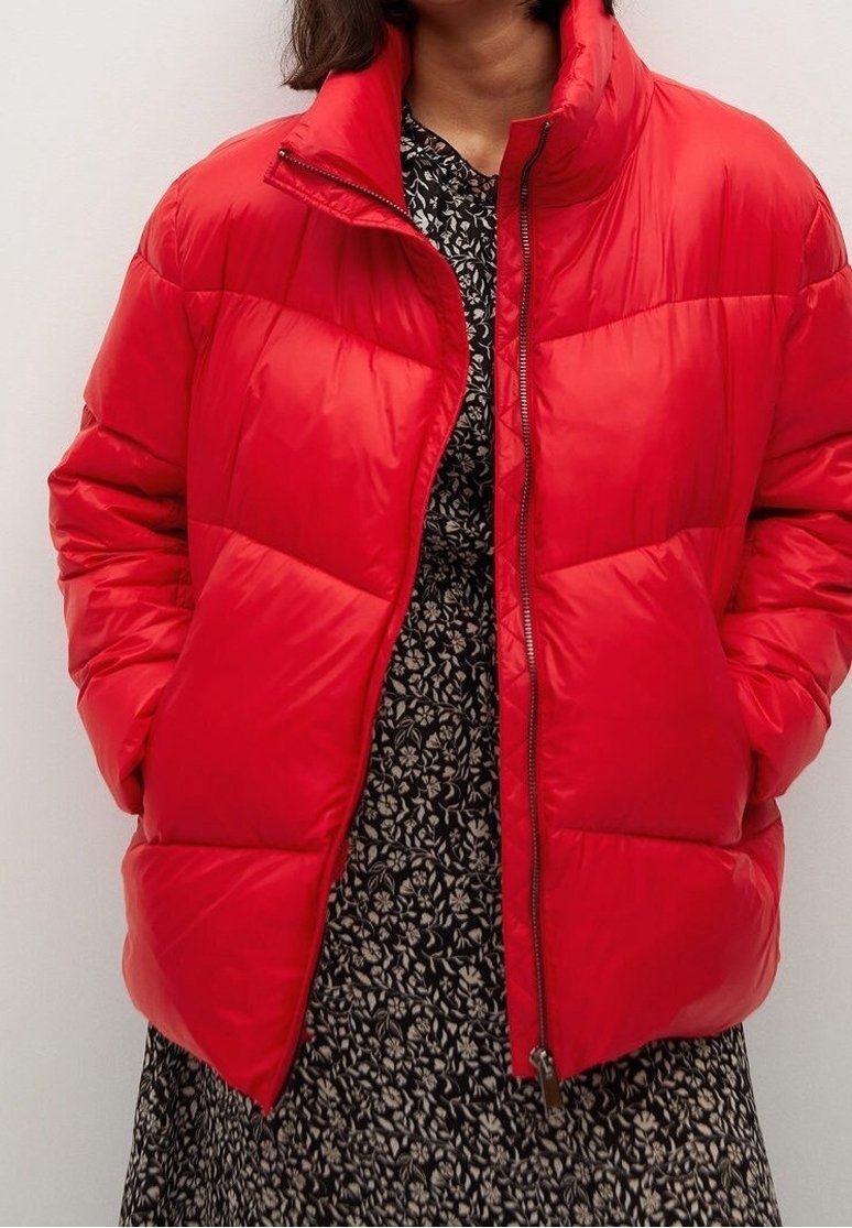 Personne portant une doudoune rouge vif sur une robe noire avec des motifs floraux blancs, les mains dans les poches de la veste, debout devant un fond uni.