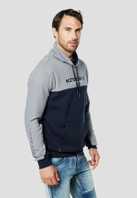 Sudadera con capucha de pulóver en azul marino y gris con medio cierre, textura a rayas en las mangas, bolsillo delantero y logo en el pecho. Material mezcla de algodón.