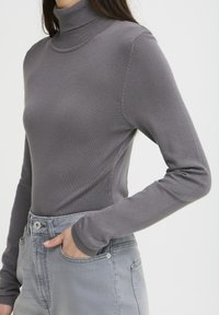 Pelēks pieguļošs augsta apkakle džemperis no ribota adījuma, ar garām piedurknēm un gludu tekstūru, komplektā ar augsto jostasvietu pelēkiem džinsiem.