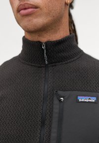 Giacca fleece Patagonia, grigia scura, con un motivo a zigzag testurizzato, colletto alto e una tasca sul petto con zip e logo.