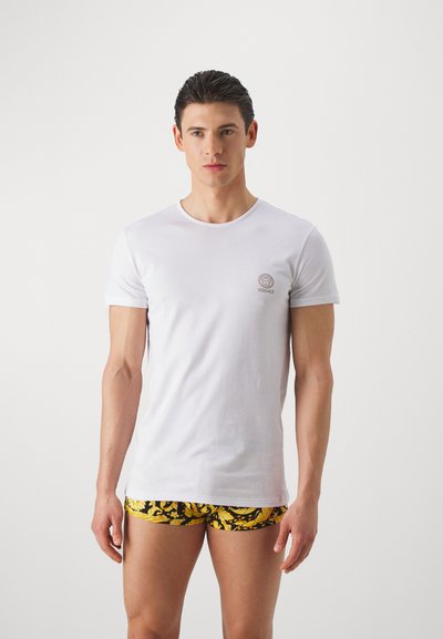 Biela bavlnená tričko s krátkymi rukávmi, na hrudi s malým zlatým logom Versace. V páre s čierno-žltými vzorovanými šortkami.