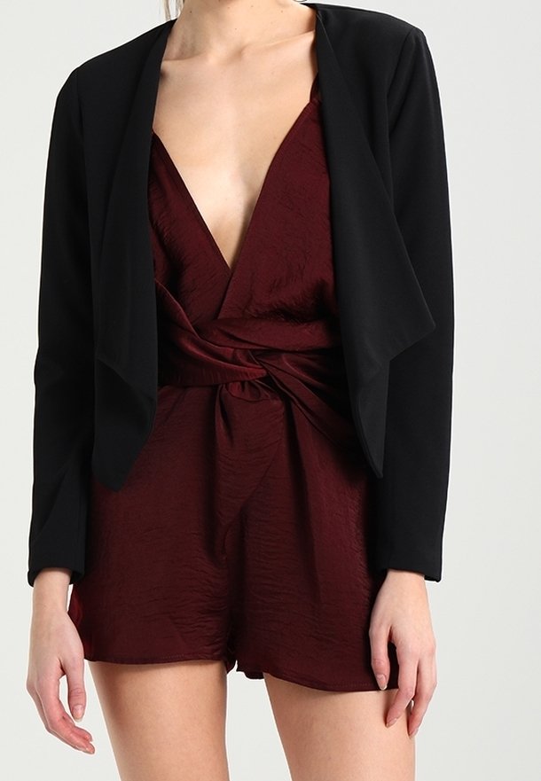 Femme portant une combinaison short style cache-cœur bordeaux foncé avec un blazer noir court ouvert à l'avant, posant devant un fond uni.