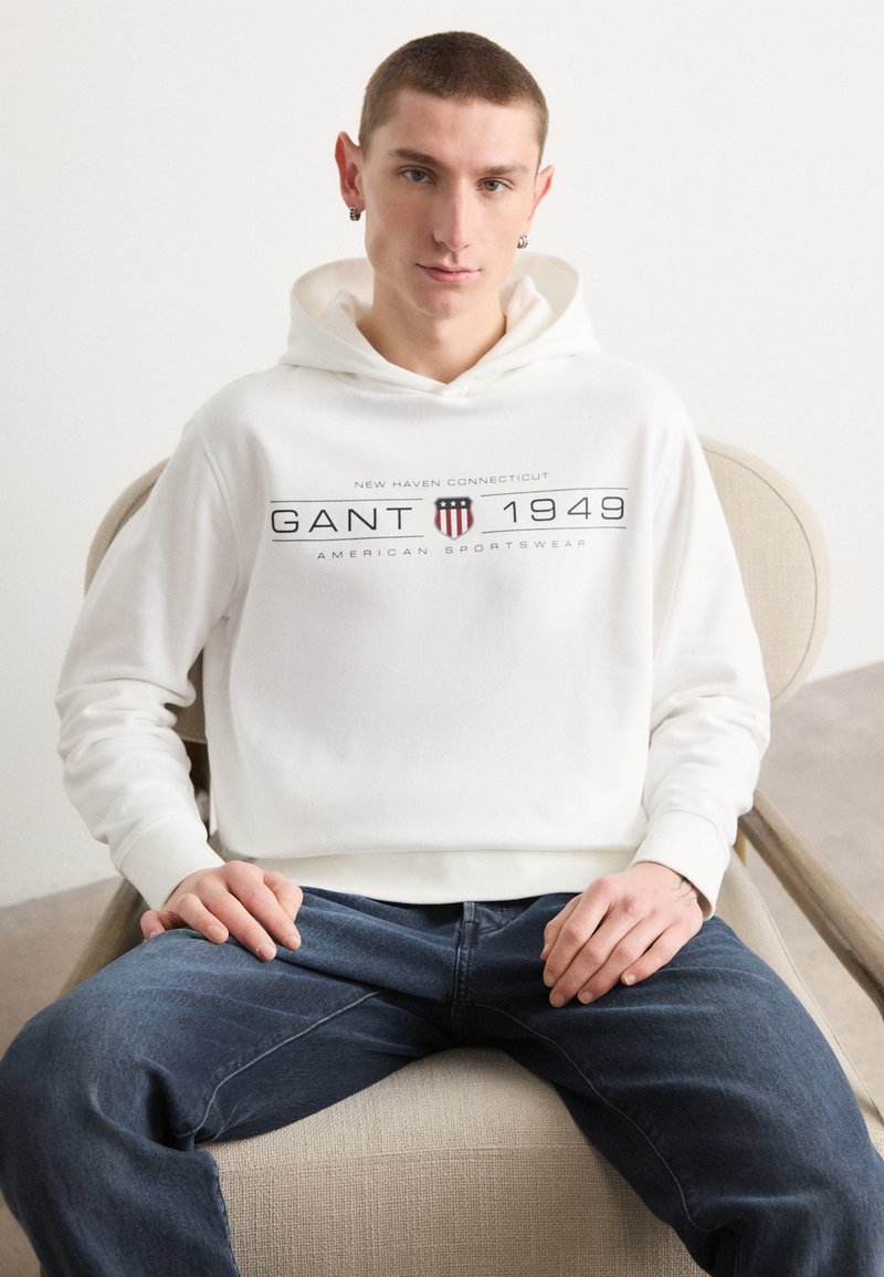 Felpa bianca con stampa frontale che presenta "GANT 1949", abbinata a jeans blu. Tessuto liscio, morbida consistenza e vestibilità rilassata.
