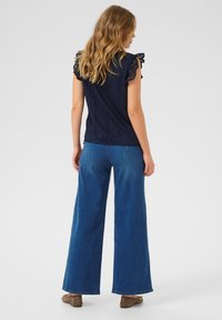 Haut bleu marine à motifs avec des manches en volants décoratives, associé à un jean bleu à pattes larges et des chaussures marron. Tissu doux et coupe décontractée.