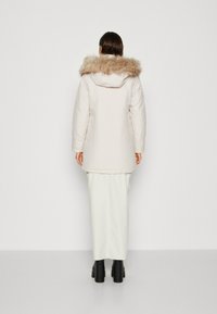 Parka beige clair avec un col en fausse fourrure, manches longues et coupe droite. Portée par-dessus une jupe blanche, des bottes noires épaisses sont visibles.