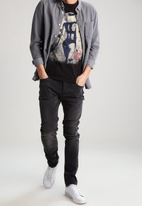 Grå knapp-up skjorta över en svart grafisk t-shirt, kombinerad med svarta jeans och vita sneakers. Enkelt design, avslappnad passform, blandade texturer.