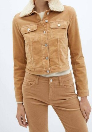 Kobieta w beżowej sztruksowej kurtce z białym kołnierzem z podszewką z owczej skóry oraz dopasowanych spodniach z wysokim stanem, stojąca na jednolitym białym tle.