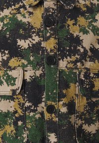 Camojacke aus Denim mit einer Mischung aus grünen, gelben und schwarzen Mustern. Verfügt über schwarze Knöpfe und Brusttaschen.