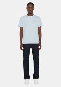 Maglietta di cotone azzurro chiaro con collo a giro e piccolo logo rosso sul petto, abbinata a jeans di denim scuro e sneaker bianche.