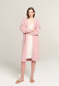 Hellrosa Strickjacke aus weichem, strukturiertem Material mit langen Ärmeln und offenem Vorderteil, über einem cremefarbenen Nachthemd und flachen Sandalen getragen.
