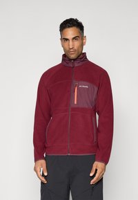 Man wearing a maroon zip-up fleece jacket with a high collar and orange zipper on the chest pocket, paired with black pants.