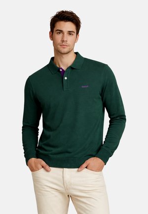 Junger Mann trägt ein dunkelgrünes langärmliges Polohemd mit lila Logo und beige Hose, steht mit den Händen in den Taschen vor einfachem Hintergrund.