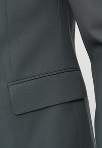 Στενή λήψη από ύφασμα σκούρου πράσινου με υφή που δείχνει τη λεπτομέρεια της τσέπης ενός ραμμένου σακακιού, καθαρές γραμμές και λεπτό φινίρισμα.