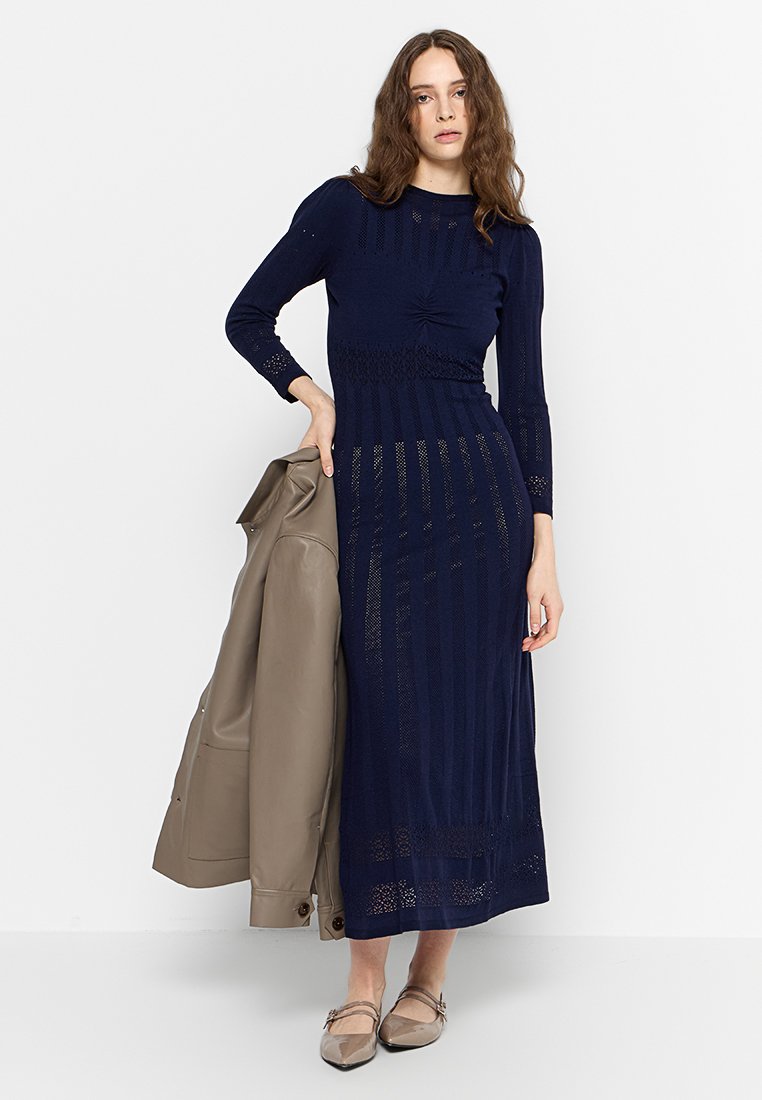 Robe en tricot ajustée de couleur marine avec des manches longues, des rayures texturées et une taille froncée. Le modèle tient une veste en cuir beige. Chaussures plates beiges.