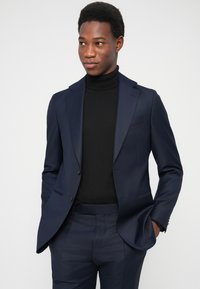 Marineblauer Anzug mit maßgeschneiderter Passform, flachen Revers und dezentem Glanz. Über einem schwarzen Rollkragenpullover getragen; glatter Stoff und strukturierte Form.