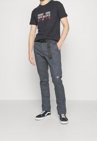 Szare spodnie o prostym kroju, czarna koszulka z grafiką z napisem "NEW BALANCE" oraz czarne sneakersy z białymi podeszwami. Widoczny detal w postaci czarnego pasa.