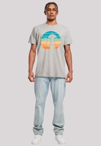 Graues T-Shirt mit kurzen Ärmeln, das ein buntes Alien-Design mit horizontalen Streifen in Blau, Grün und Orange zeigt, kombiniert mit hellblauen Jeans und weißen Sneakern.