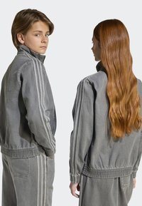 Veste en denim gris avec col montant, taille élastique et accents rayés sur les manches ; portée avec un pantalon gris assorti.