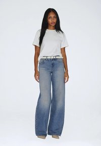 Femme debout portant une chemise blanche à manches courtes, un jean bleu à jambes larges avec une ceinture blanche, et des chaussures beiges pointues sur un fond clair uni.