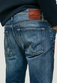 Jean en denim bleu avec une finition délavée, des coutures contrastantes et une étiquette en cuir marron avec le logo "Pepe Jeans" sur la ceinture.
