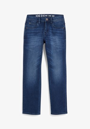 Barn iført farveblok-hættetrøje, blå cowboybukser med opbuk, hvide sneakers, stående med den ene hånd i lommen mod en ensfarvet baggrund.